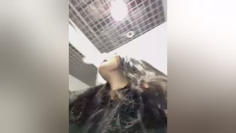 Video of a woman in a dark coat, her hair disheveled, standing in a shower with a grid-tiled ceiling and white walls, water splashing.