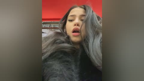 Media: A video of a young woman with long, silver hair and light skin, wearing a black fur coat, captured indoors with a red wall and metallic window blinds in the background.