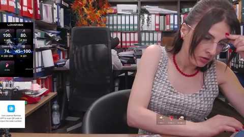 Media: Video of a Caucasian woman with brown hair, wearing a patterned top and red necklace, sitting at a desk, looking tired. Background shows a messy office with books, papers, and a black chair.