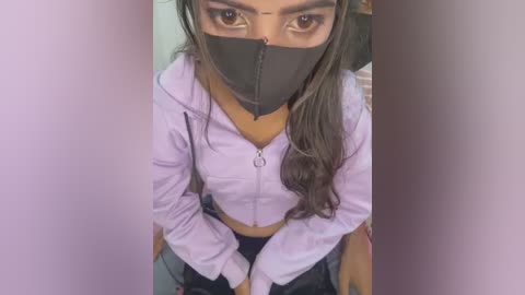 Media: Video of a young woman with medium skin tone, wearing a black face mask, lavender hoodie, and jeans, taken from an elevated angle.