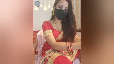 Media: A video of a South Asian woman in a red and gold sari with gold embroidery, wearing a black face mask, gold bangles, and a necklace, seated on a bed with a pink blanket.