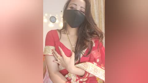 Media: Video of a South Asian woman wearing a red sari with gold embroidery, black mask, and black gloves, covering her chest, in a softly lit room.