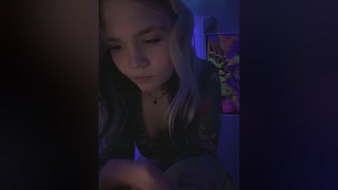 Media: A dimly-lit video shows a young woman with long, wavy, platinum blonde hair, wearing a dark, lacy top, and a necklace, concentrating on a smartphone screen.