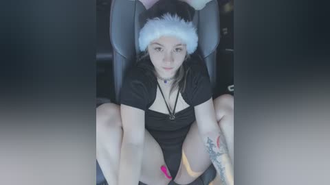 Media: Video of a young, light-skinned woman with brown hair, wearing a Santa hat, black dress, and pink nail polish, sitting in a grey gaming chair, legs crossed, with tattoos on her thighs.