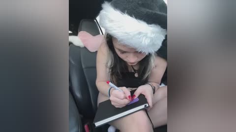 Media: Video of a young girl with long blonde hair wearing a black and white Santa hat, seated in a car, writing in a notebook.