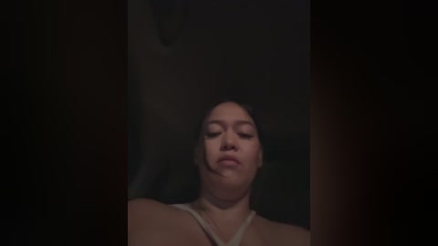 Media: A dimly lit video features a young Asian woman with straight black hair and light skin, wearing a white top, with her eyes closed and mouth slightly open, suggesting a contemplative or emotional state. The background is dark and indistinct.