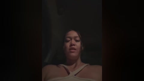 Media: Video of a young woman with light brown skin and medium-length, dark hair, wearing a beige tank top, looking directly at the camera with a neutral expression. The background is dark and indistinct.