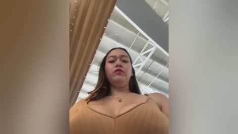 Media: Video of a young woman with light brown skin, wearing a tan spaghetti-strap top, standing in a modern, industrial space with white beams and beige curtains.