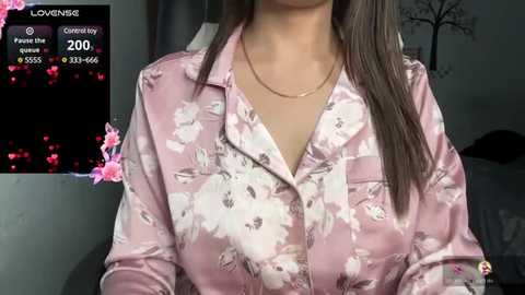 Media: Video of a woman with medium skin tone, long brown hair, wearing a pink floral blouse, thin gold necklace, in a dimly lit room with pink cherry blossom decor.