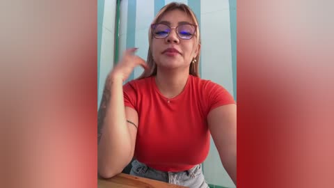 Media: Video of a young Asian woman with light skin, short brown hair, and glasses, wearing a red T-shirt and blue jeans, standing in a bathroom with a blue and white striped curtain.