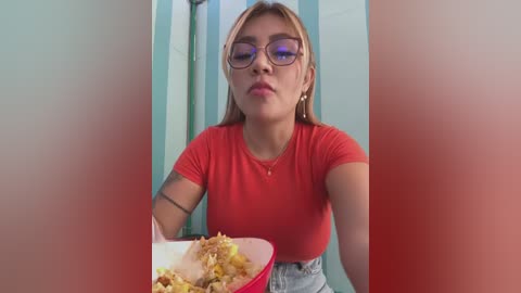 Media: Video of a young woman with light skin, long blonde hair, wearing glasses, a red top, and jeans, eating cereal from a bowl in a colorful, tiled bathroom.