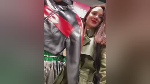 Media: Video of a woman with shoulder-length brown hair, wearing a green trench coat, standing next to a mannequin in a shiny, black leather jacket with a red panel.