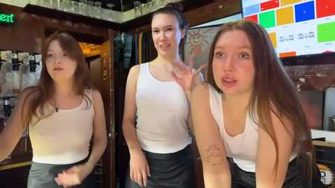 Media: Video of three young women in a bar, one pointing, wearing white tank tops and black skirts, wooden background with TV screens.