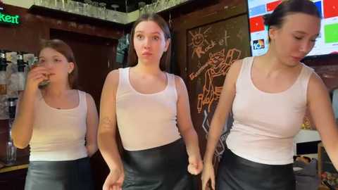 Media: Video of three young women in white tank tops and black skirts, standing side-by-side in a dimly lit bar with chalkboard art, illuminated screens, and bar stools.