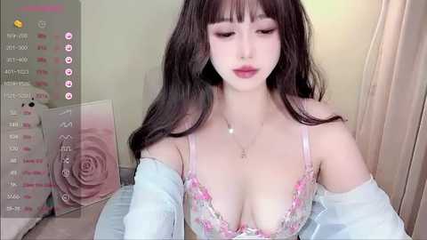 Media: Video of an Asian woman with long black hair and fair skin, wearing a pink lace bra and off-shoulder top, in a room with a floral wall decor.