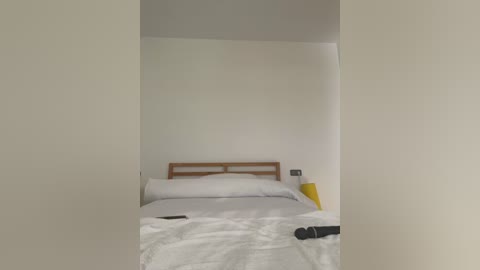 Media: Video of a minimalist, unmade bed with white sheets and a wooden headboard against a plain white wall, with a yellow lamp and a black remote control on the nightstand.