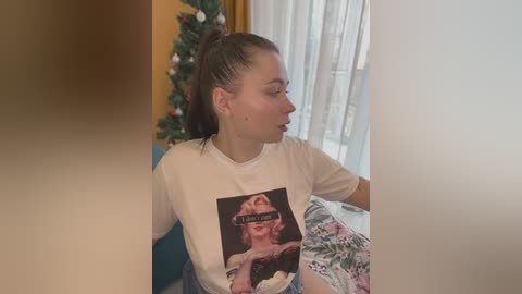 Media: Video of a fair-skinned, brunette woman with a ponytail, wearing a Marilyn Monroe t-shirt, sitting on a couch beside a decorated Christmas tree.