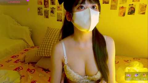 Media: Video of an East Asian woman with long black hair and a white mask, wearing a floral-patterned bra, lying on a yellow bed with a checkered pillow. The room is yellow with photos on the wall.