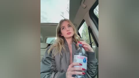 Video of a blonde woman in a gray jacket holding a water bottle, seated in a car with a sky and trees visible through the window.