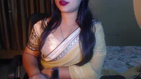 Media: Video of a South Asian woman with long dark hair, wearing a yellow sari with intricate silver embroidery and a white lace blouse, sitting indoors at a desk.