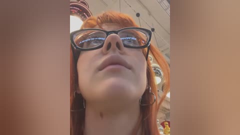 Media: A close-up video of a young woman with fair skin and long, bright orange hair, wearing black-rimmed glasses and a black dress. The background shows a mall with a ceiling, signs, and hanging lights.