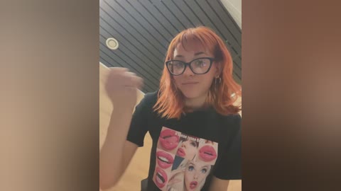 Media: Video of a fair-skinned woman with red hair, wearing black-framed glasses, a black t-shirt with a graphic of red lips, and a beige background with a textured ceiling.