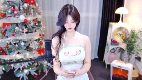 Media: Video of an East Asian woman with long black hair, wearing a white apron with hearts, standing in a brightly lit living room with a decorated Christmas tree and modern furniture.