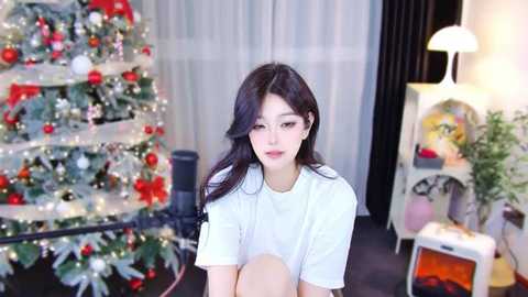 Media: Video of a young East Asian woman with long dark hair, wearing a white t-shirt, seated in a cozy living room with a decorated Christmas tree and warm lighting.