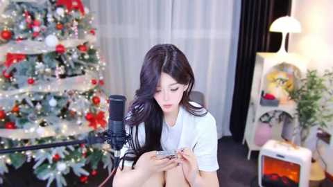 Media: A video of an Asian woman with long black hair, wearing a white shirt, sitting at a desk, looking at a smartphone, in a room with a decorated Christmas tree and a desk lamp.