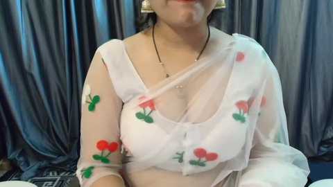 Media: Video of a South Asian woman with medium skin tone wearing a sheer white blouse adorned with cherry print, showcasing medium-sized breasts, against a dark, draped curtain background.