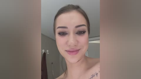 Media: Video of a smiling, light-skinned woman with brown hair, wearing makeup, including dark eyeliner and mascara. She has a tattoo on her shoulder. The background is a neutral-toned bathroom with towels and a door.