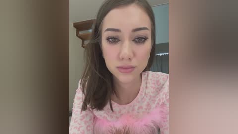 Media: Video of a young woman with light skin, straight brown hair, and light makeup, wearing a pink floral-patterned shirt. Background shows a wooden door and blurred interior.