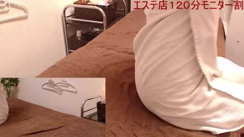 Media: Video of a neatly made bed with a white pillow, beige blanket, and a metallic coat hanger on a white wall. Japanese text overlays the image.