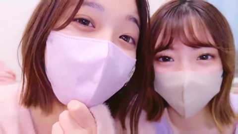 Media: Video of two young East Asian women with straight brown hair, wearing light-colored face masks, smiling closely. They are indoors, with a soft, blurred background.