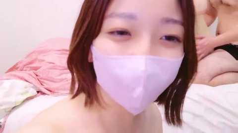 Media: Video of an Asian woman with shoulder-length brown hair, wearing a white face mask, lying on a bed with pink sheets, blurred background.