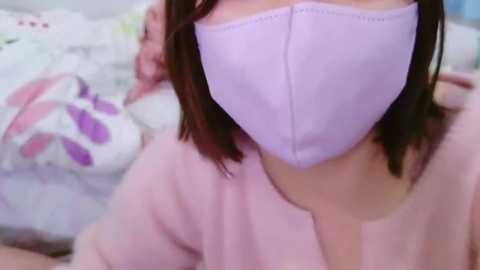 Media: Video of a person wearing a lavender surgical mask, pink top, and white pillow with floral design in the background.