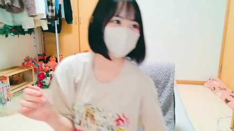 Media: Video of a young East Asian woman with a bob haircut, wearing a floral t-shirt, face mask, and black pants, sitting in a cluttered room with clothes, toys, and a desk.