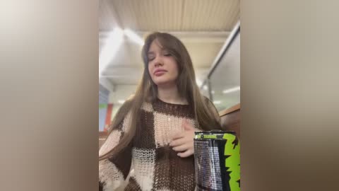 Media: Video of a young girl with long brown hair, wearing a chunky, patchwork knit sweater, holding a small, green and black electronic device, standing in a brightly lit, modern indoor setting with white ceilings and recessed lights.