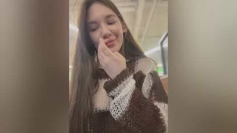 Media: Video of a young woman with long, straight brown hair, wearing a patterned brown sweater, standing in an indoor space with beige walls and a ceiling. She has her finger to her lips in a thoughtful gesture.