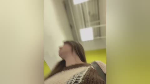 Media: A blurred video of a young woman with long brown hair, wearing a brown and beige knitted sweater, leaning against a yellow wall in a brightly lit room.