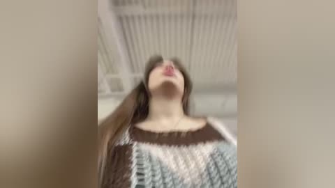 Media: A blurry video of a woman with long brown hair, wearing a brown and white knitted sweater, standing in a white room with visible air conditioning vents.