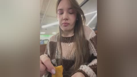 Media: Video of a young woman with long brown hair, wearing a beige and brown knitted sweater, eating a banana on a bus, with blurred background.