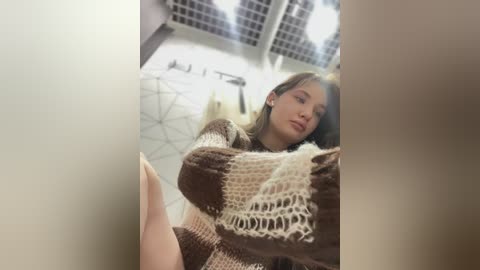 Media: Video of a young woman with straight brown hair, wearing a brown and beige knitted sweater, leaning against a white wall with geometric patterns, in a modern, brightly lit bathroom.