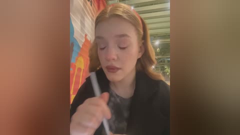 Media: Video of a young girl with fair skin and light brown hair, wearing a black coat, holding a pen, standing in front of a colorful mural.