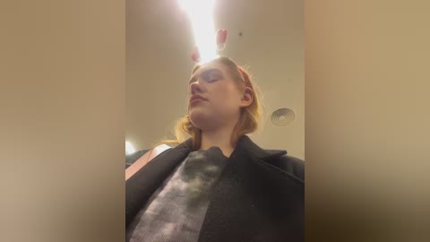 Media: Video of a young woman with fair skin, blonde hair, and a red headband, wearing a black coat, looking up at a ceiling light in a beige-walled room.