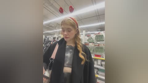 Media: Video of a young blonde woman with a red headband, wearing a black coat and gray crop top, shopping in a bright, organized store with shelves filled with colorful products.