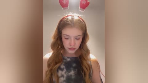 Media: Video of a young woman with long, wavy auburn hair wearing a red headband with white snowflakes, standing in front of a beige wall with red, Christmas-themed ornaments hanging above her head.