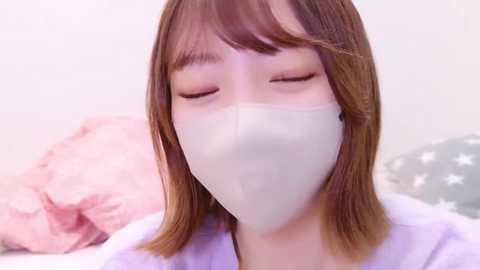 Media: Video of a young Asian woman with shoulder-length light brown hair, wearing a white face mask and a light purple shirt, eyes closed, sitting on a bed with pink and star-patterned pillows.