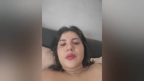 Media: Video of a Latina woman with medium skin tone, dark hair, and full lips, lying topless on a black couch, her arms behind her head. The background is blurred, with a white wall.