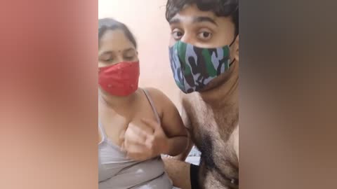 Media: A video of two people wearing face masks in a dimly lit room. The woman, with medium skin tone and black hair, is pulling down her gray tank top, exposing her breast. The man, with medium skin tone and dark hair, has a surprised expression.